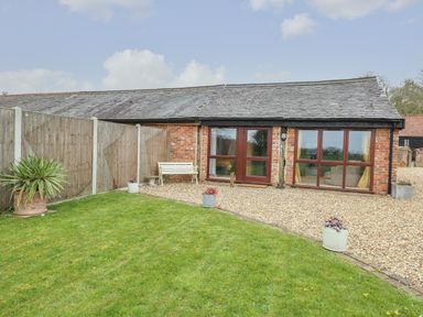 A backyard with grass, potted plants, a bench, gravel area, and a brick building with wooden doors and windows at Bee Happy Barn in Beighton near Acle