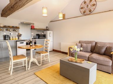 A living area with a round dining table and two chairs a sofa with cushions and a coffee table with flowers and kitchen appliances at Bee Happy Barn in Beighton near Acle