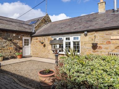A courtyard with gravel and potted plants in front of a stone house with white windows and a door at West Farm Cottage in Longhorsley