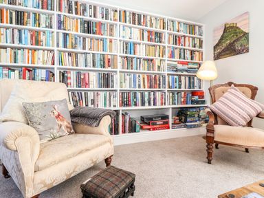 A reading room with a large bookshelf filled with books two armchairs a lamp and a small footstool at West Farm Cottage in Longhorsley