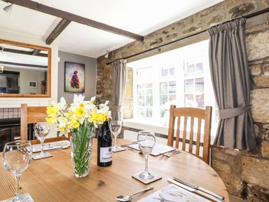 A dining table set with wine glasses and a vase of flowers near a window with curtains and stone walls at West Farm Cottage in Longhorsley