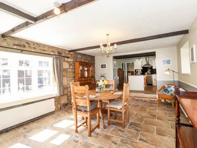A dining area with a wooden round table and chairs next to a stone wall and window at West Farm Cottage in Longhorsley