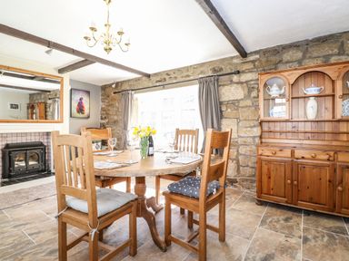 A dining room with a round wooden table set with glasses and cutlery four wooden chairs a wooden cabinet and a fireplace at West Farm Cottage in Longhorsley