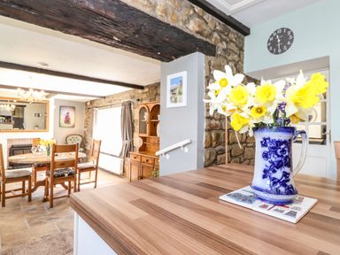A kitchen counter with a floral vase and dining table with chairs in a room with stone walls and wooden beams at West Farm Cottage in Longhorsley