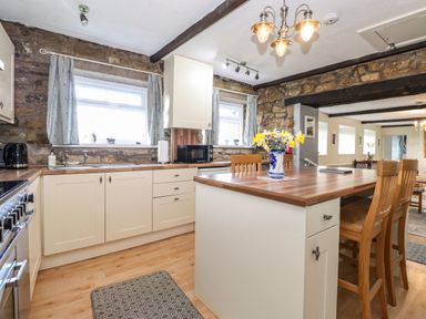 A kitchen with a wooden island and chairs stone walls and two windows with curtains at West Farm Cottage in Longhorsley