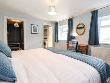 A bedroom with a bed and blue pillows two windows with blue curtains a wooden desk with a chair and a round mirror at West Farm Cottage in Longhorsley
