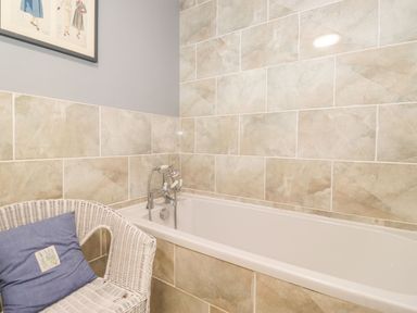 A bathroom with a white bathtub with tiled walls and a white wicker chair with a blue cushion at West Farm Cottage in Longhorsley