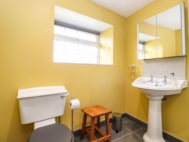 A bathroom with a toilet, wooden stool, trash bin, sink with mirror cabinet and window at West Farm Cottage in Longhorsley