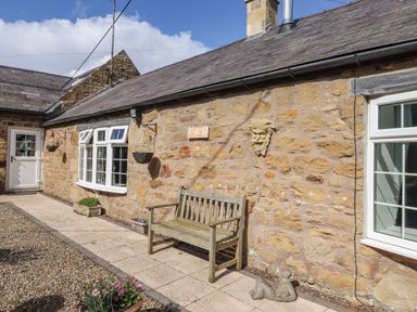 The exterior of a stone cottage with a wooden bench on a tiled path and flower pots by the walls at West Farm Cottage in Longhorsley