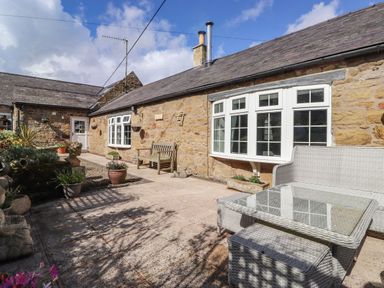 An outdoor patio area with stone walls white framed windows wooden bench and wicker seating at West Farm Cottage in Longhorsley