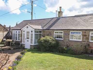 A stone cottage with white framed windows and a white door surrounded by a garden with flowers and outdoor furniture at West Farm Cottage in Longhorsley