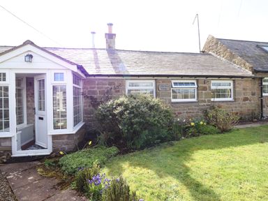 The exterior of a stone cottage with white framed windows and a garden with flowers and shrubs at West Farm Cottage in Longhorsley