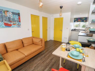 A living room with a brown sofa three yellow doors a dining table set with blue dishes and yellow mugs and a kitchen area visible at Nanny's Little House in Sandown