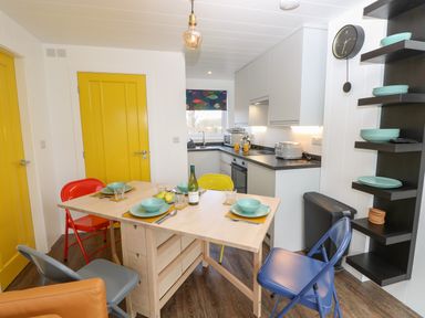 A kitchen with a wooden table set for four, colorful chairs, yellow doors, appliances, and wall shelves with plates at Nanny's Little House in Sandown