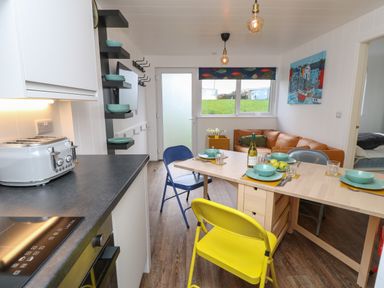 A kitchen and dining area with a toaster and induction stove a wooden table set with plates bowls and glasses yellow and blue chairs and a brown sofa near a window at Nanny's Little House in Sandown