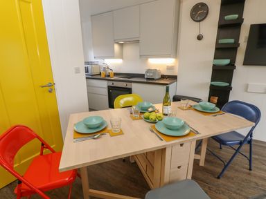 A kitchen with a wooden dining table set with bowls plates glasses and cutlery and colorful chairs at Nanny's Little House in Sandown