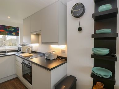 A kitchen with white cabinets a black countertop a sink a toaster a microwave a wall clock and shelves with green plates at Nanny's Little House in Sandown