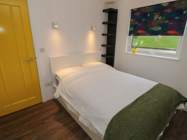 A bedroom with a double bed a yellow door a window with a fish patterned blind and a black corner shelf at Nanny's Little House in Sandown