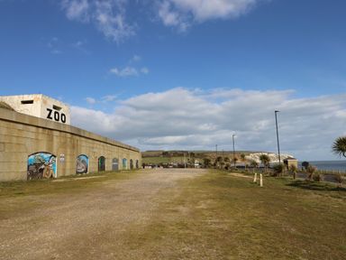 An outdoor area with a stone wall painted with murals and a building labeled zoo near a road and coastline at Nanny's Little House in Sandown