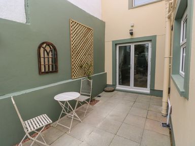 An outdoor patio area with two white chairs and a small round table near a green wall with a mirror and trellis at Stones Throw in Tenby