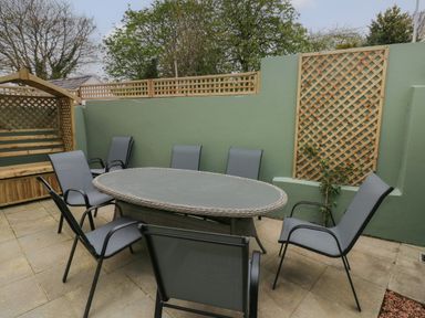 An outdoor patio with a round table surrounded by six chairs and a wooden bench with lattice against a green wall at Stones Throw in Tenby
