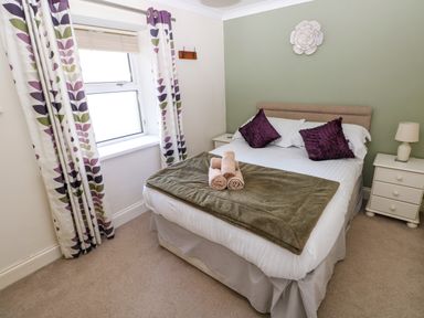 A bedroom with a double bed with purple pillows and rolled towels on a green blanket a window with patterned curtains and a bedside table with a lamp at Stones Throw Tenby