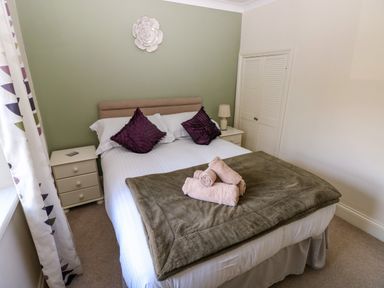 A bedroom with a double bed, two purple pillows, rolled towels on a blanket, nightstands with a lamp and drawers, and a white floral wall decoration at Stones Throw in Tenby