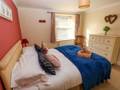 A bedroom with a bed featuring multiple pillows and folded towels a wooden chair a white dresser with a vase of flowers and a window with striped curtains at Stones Throw in Tenby