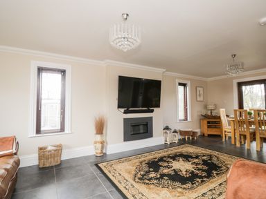 A living room with a wall-mounted TV a fireplace a patterned rug and a dining table with chairs at Mill End in Eamont Bridge near Penrith