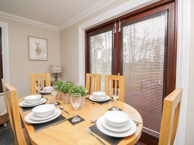 A dining room with a wooden table set for six with white dishes and plants near wooden framed glass doors with blinds at Mill End in Eamont Bridge near Penrith