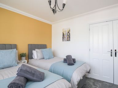 A bedroom with two single beds with blue and gray bedding and towels on each bed a yellow accent wall and a white wall with a painting and double white doors at Mill End in Eamont Bridge near Penrith