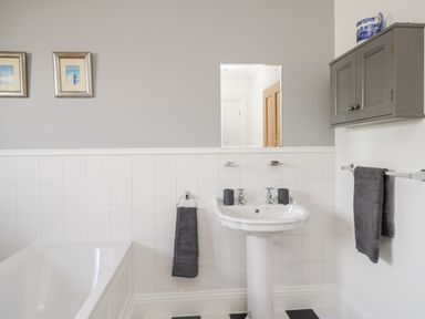 A bathroom with a white bathtub a pedestal sink two gray towels a wall cabinet and two framed pictures at Mill End in Eamont Bridge near Penrith