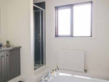 A bathroom with a bathtub, a window with blinds, a radiator, a shower with glass door, and a cabinet with a plant on top at Mill End in Eamont Bridge near Penrith