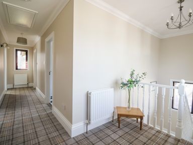A hallway with beige walls a plaid carpet a small wooden table with flowers and white railing at Mill End in Eamont Bridge near Penrith