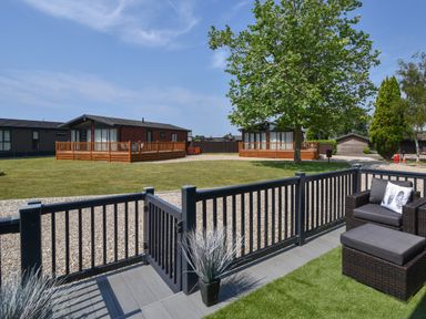 A fenced patio area with a chair and ottoman overlooking wooden lodges and a tree at Oakwood 2 Pinewood Retreat in Pinewood