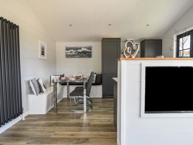 A dining area with glass table and built-in seating with cushions next to a kitchen with dark cabinets and a wall-mounted TV at Oakwood 2 @ Pinewood Retreat in Pinewood