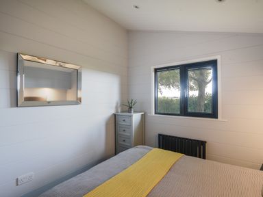 A bedroom with a bed with gray and yellow bedding a window a small cabinet with a plant and a wall mirror at Oakwood 2 @ Pinewood Retreat in Pinewood