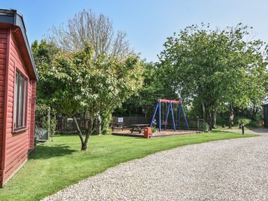 A red cabin beside a gravel path with a tree and a playground with swings and picnic benches at Oakwood 2 at Pinewood Retreat in Pinewood