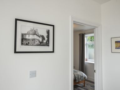 A bedroom visible through a doorway with a bed and a window with curtains and framed pictures on the walls at Lyme Zest in Lyme Regis