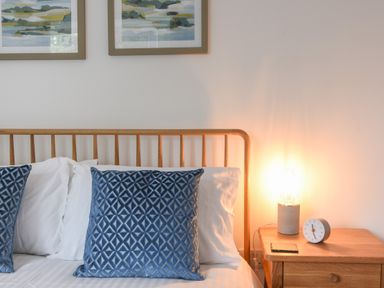 A bed with white sheets and blue pillows next to a wooden nightstand with a lit lamp and a clock at Lyme Zest Lyme Regis