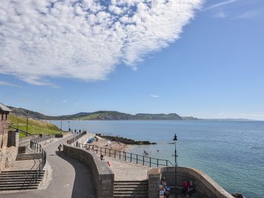 A coastal walkway with people sitting and walking near the sea with hills in the background at Lyme Zest in Lyme Regis