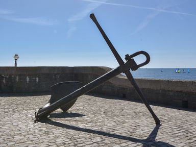 A large metal anchor on a cobblestone surface near a sea wall with sailboats on the water at Lyme Zest in Lyme Regis
