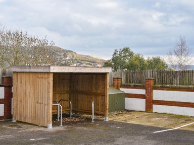 A wooden bike shelter with metal bike racks beside a fence with trees and hills in the background at Lyme Zest in Lyme Regis