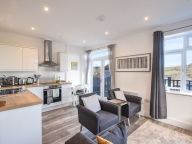 A kitchen and living area with two armchairs a small wooden table and curtains on the windows at Lyme Zest Lyme Regis