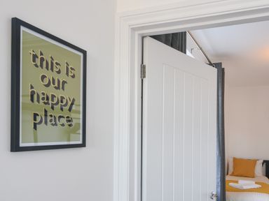 A framed sign on a wall next to an open door leading to a bedroom with a bed with mustard yellow pillows and a blanket at Lyme Zest in Lyme Regis