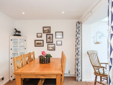 A dining room with a wooden table and chairs a white cabinet with glassware black and white photos on the wall and a wooden chair with a cushion near a window with patterned curtains at Neaudd Wen Red Wharf Bay near Benllech