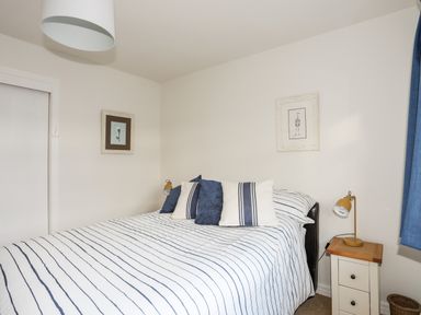 A bedroom with a bed striped bedding blue and white pillows a nightstand with a yellow lamp and framed pictures on white walls at Neaudd Wen Red Wharf Bay near Benllech