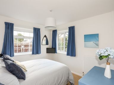 A bedroom with a bed pillows blue curtains windows a small television a radiator a blue chest and a vase with flowers at Neaudd Wen in Red Wharf Bay near Benllech