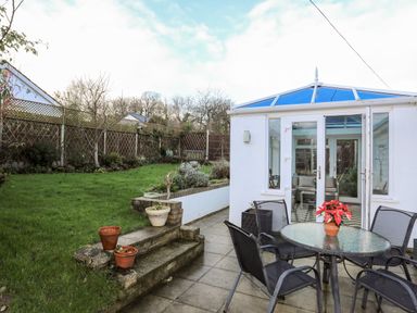 A garden with a grass lawn and a paved patio area with a glass table and four chairs outside a white conservatory at Neaudd Wen in Red Wharf Bay near Benllech