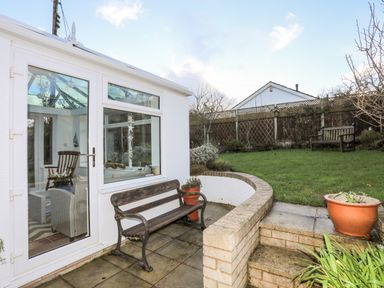A patio with a wooden bench next to a white conservatory and a garden with grass and another bench in the background at Neaudd Wen Red Wharf Bay near Benllech